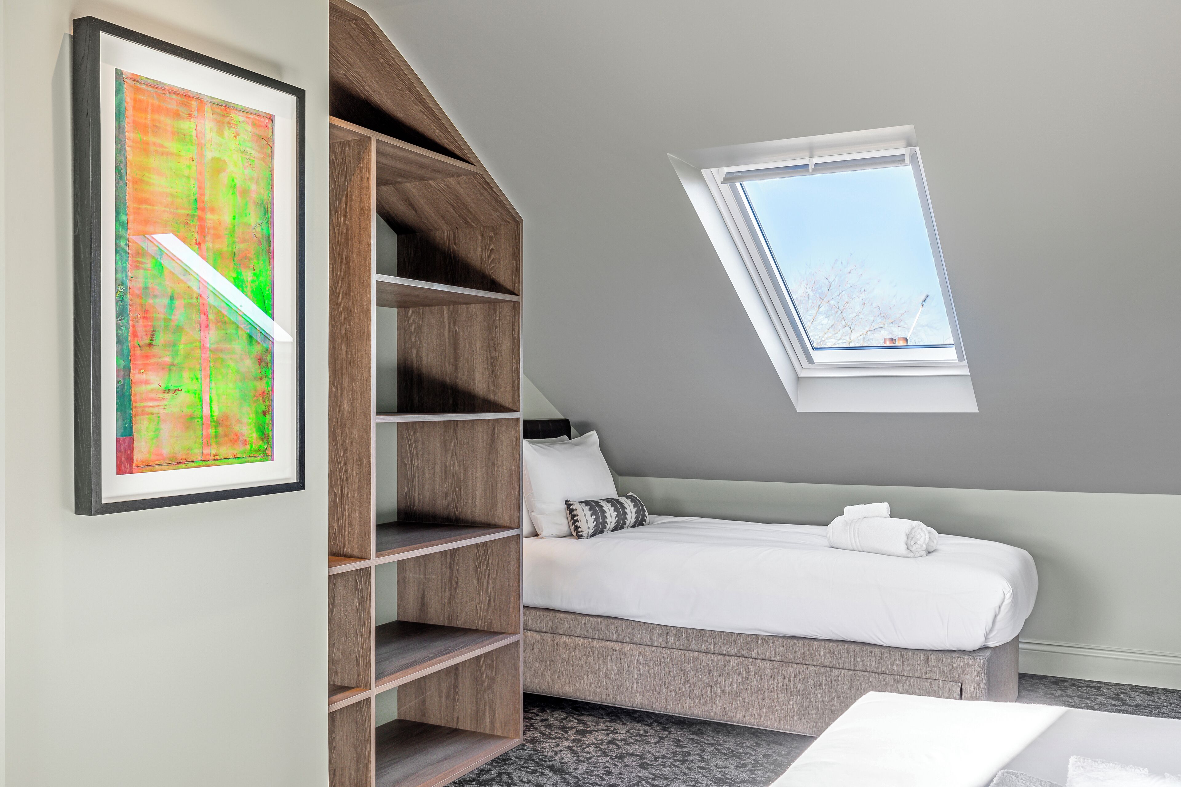 A bedroom with a sloped ceiling, a skylight, a bed with white bedding, and a wooden shelving unit.