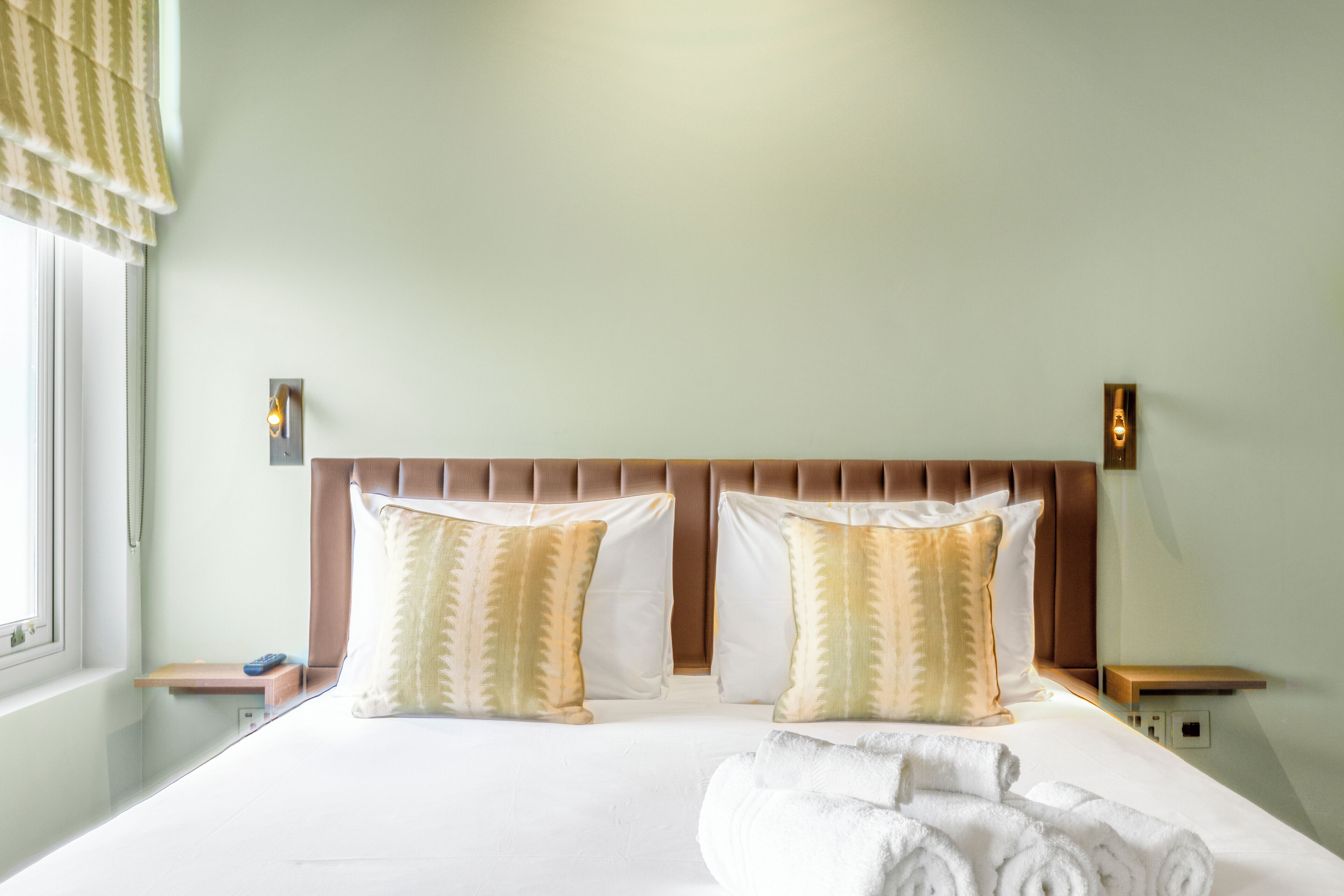 Una habitación de hotel con una cama grande, dos almohadas, cabecero, mesitas de noche y un ventanal con cortinas.