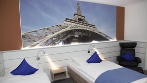 A hotel room with two beds, a large Eiffel Tower poster on the wall, a massage chair, and a small bedside table.