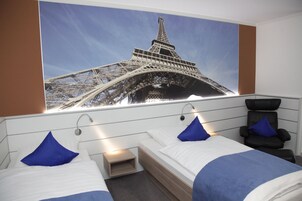 Habitación de hotel con dos camas, un cartel grande de la Torre Eiffel en la pared, una silla de masaje y una mesita de noche pequeña.