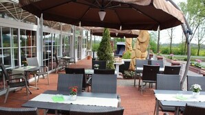 An outdoor patio area with tables and chairs, covered by umbrellas, and surrounded by potted plants and a stone sculpture.