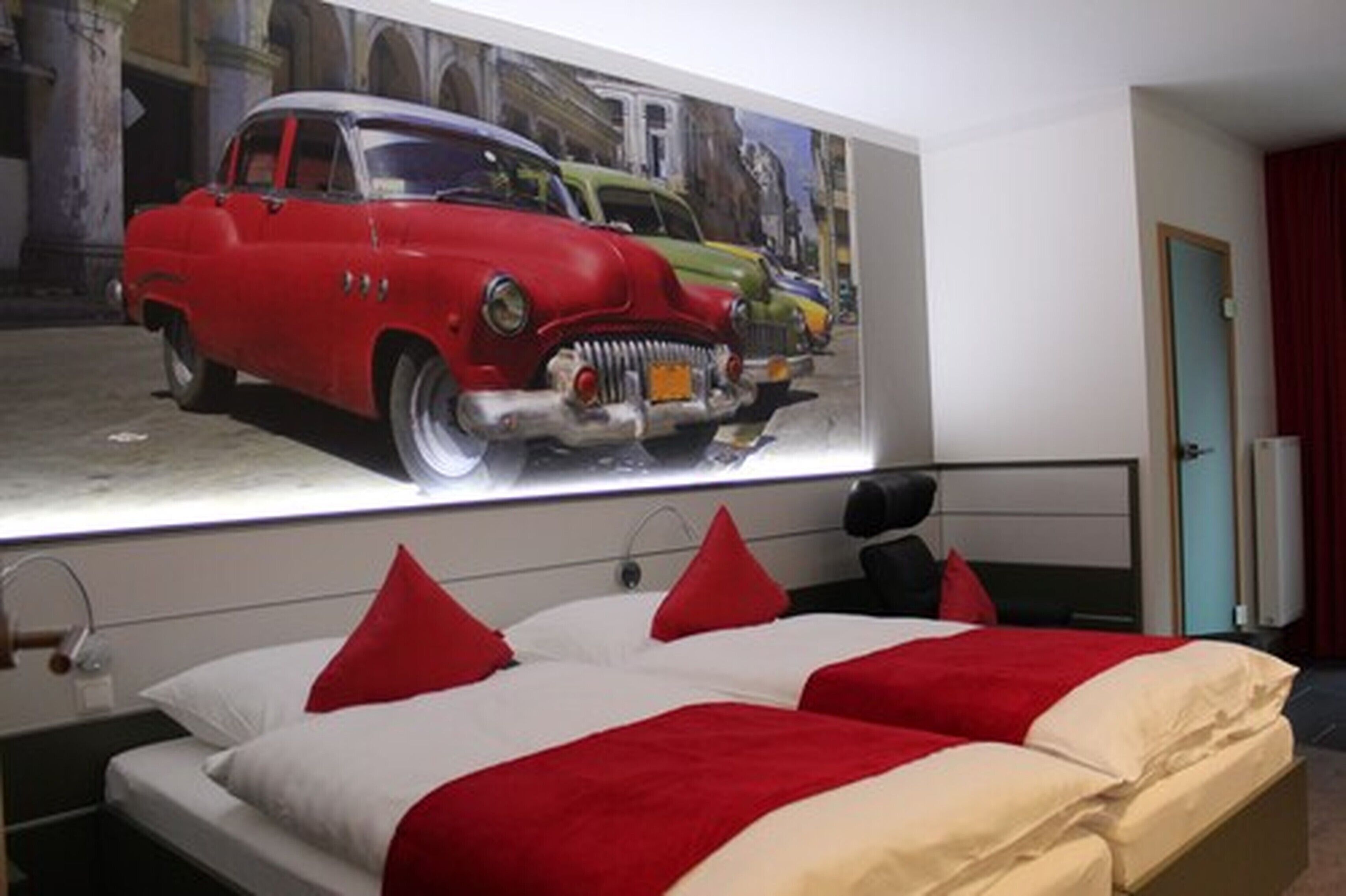 A hotel room with a large bed, red and white bedding, and a wall mural of vintage cars.