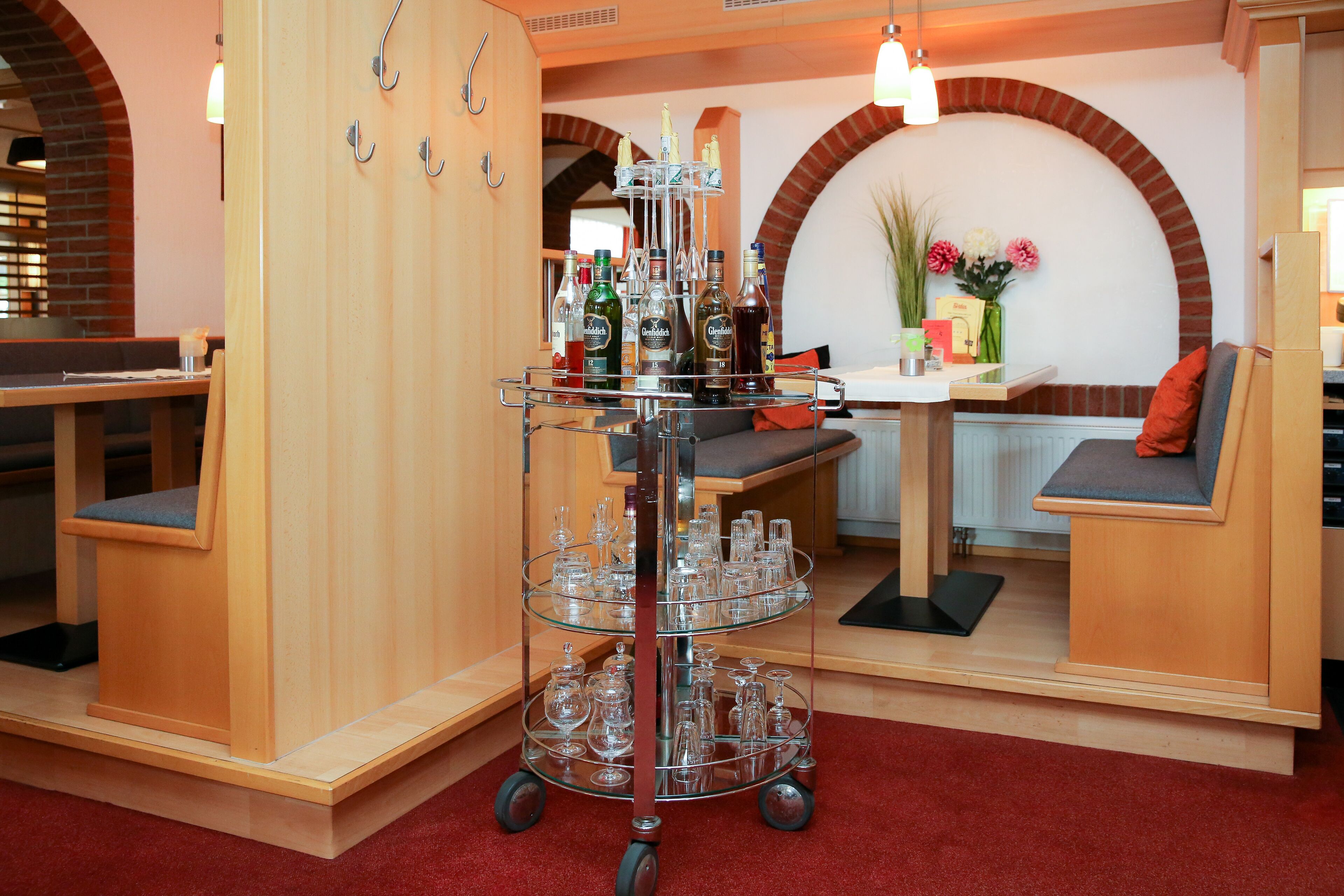 Interior de restaurante con un carro de vidrio de tres niveles lleno de botellas y copas, muebles de madera y un arco de ladrillo al fondo.