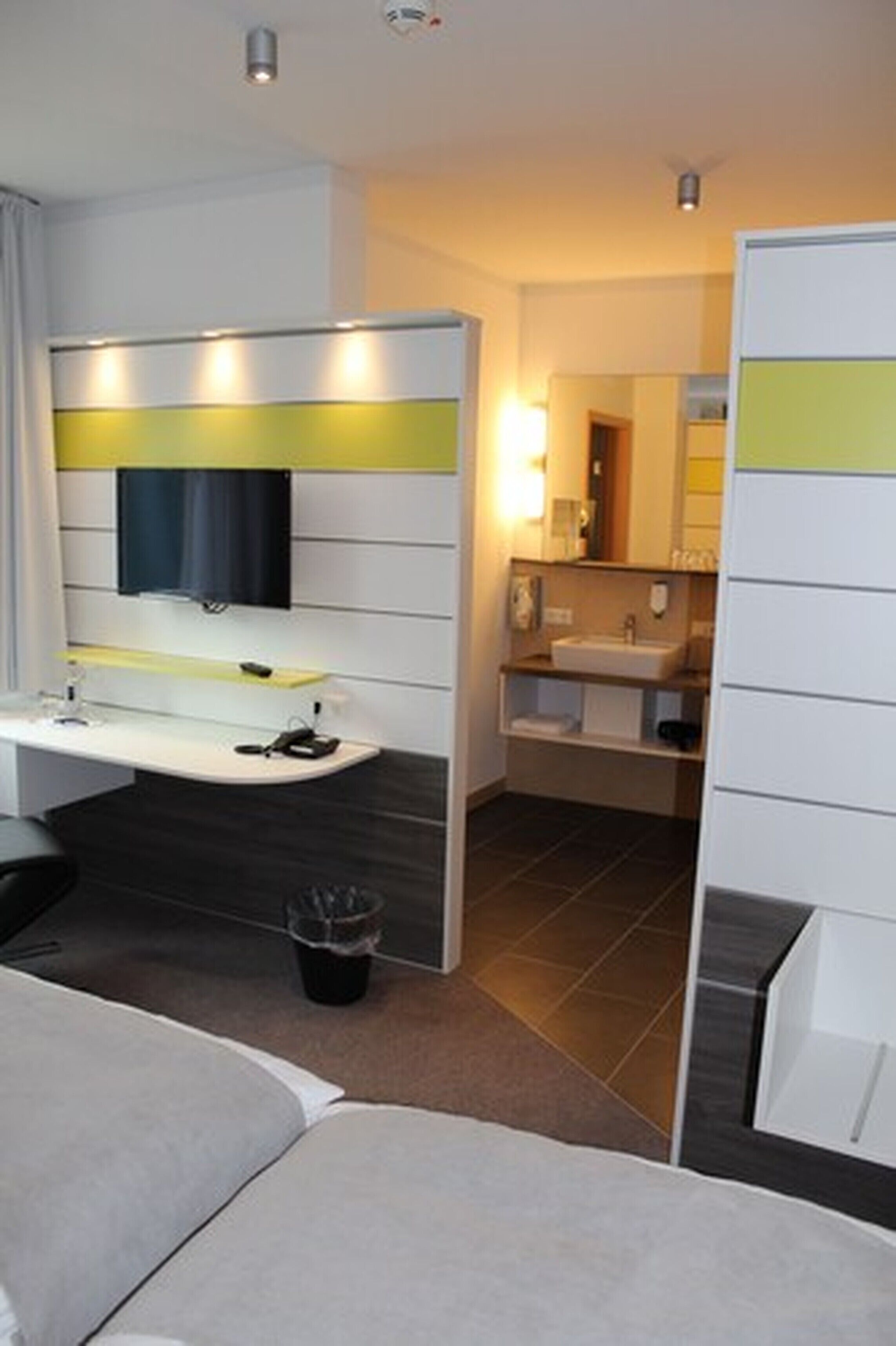 Una habitación de hotel moderna con sofá, escritorio con computadora, televisor de pantalla plana, baño con lavamanos y espejo, y un cubo de basura.