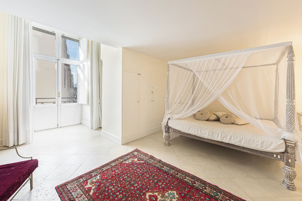 Une chambre à coucher avec un lit à baldaquin, un tapis rouge à motifs et une vue sur la ville par une grande fenêtre.