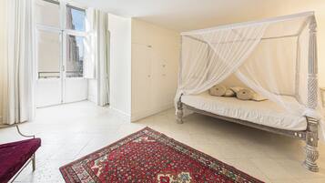 A bedroom with a four-poster bed, a red patterned rug, and a view of the city through a large window.