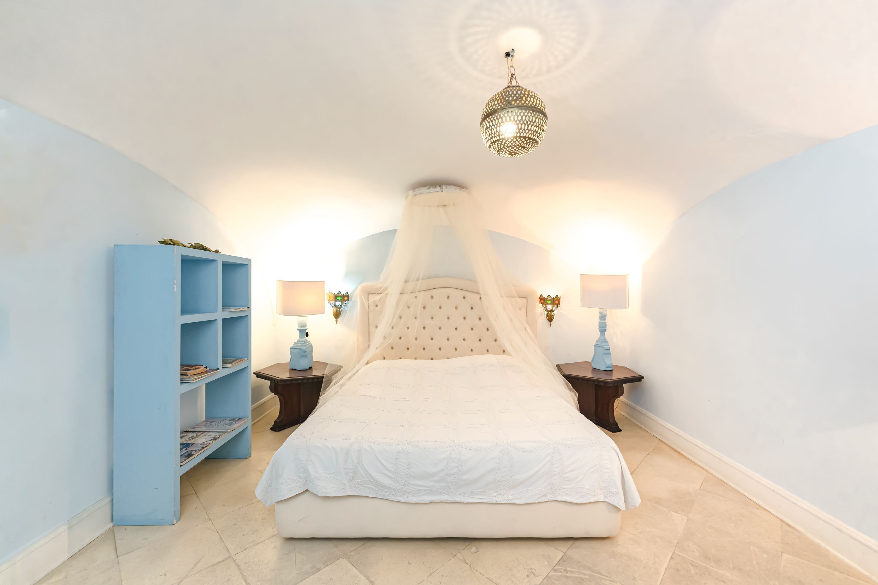 Un dormitorio con una cama, una estantería azul, dos mesitas de noche con lámparas y una lámpara decorativa en el techo.