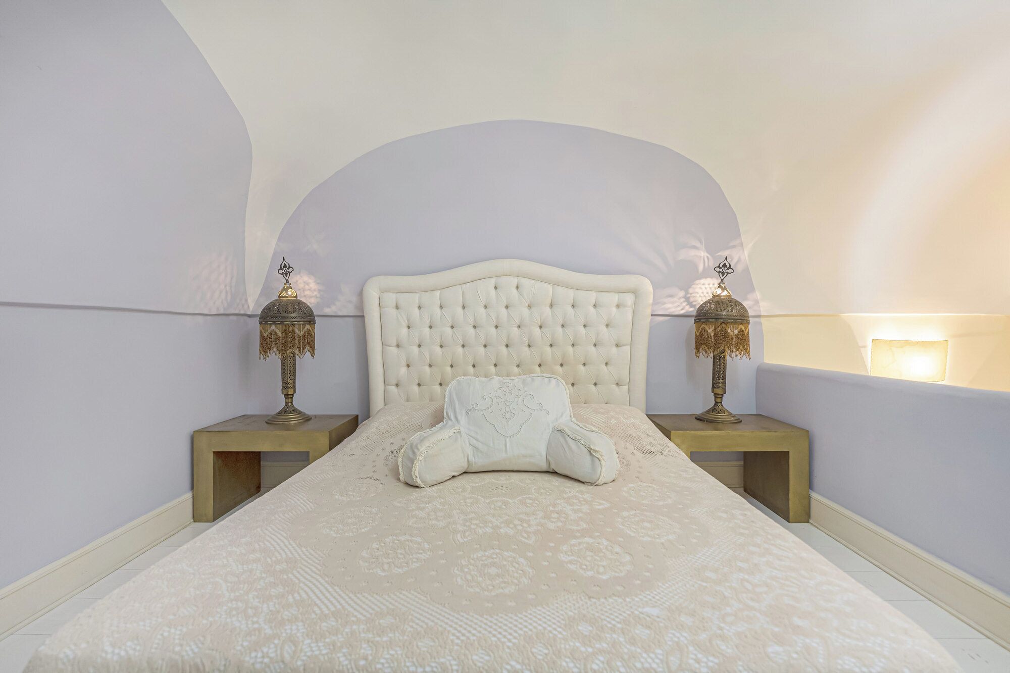 A bedroom with a tufted headboard, a patterned bedspread, two bedside tables with lamps, and a wall-mounted light.