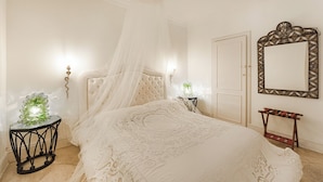 Une chambre à coucher avec un lit à baldaquin, une table ronde ornée d’un vase vert, un miroir avec un cadre décoratif et un banc avec des coussins rouges.