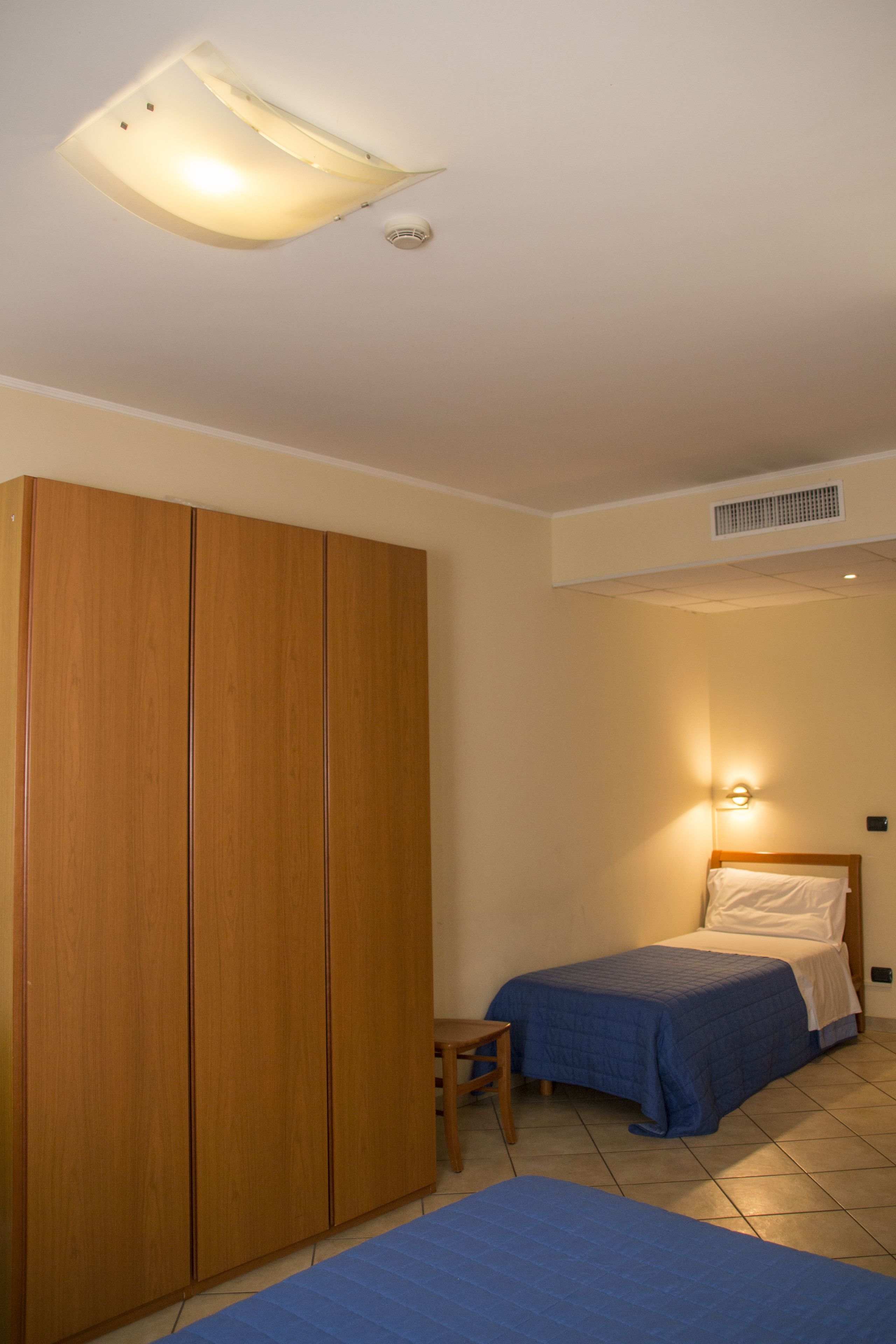 A bedroom with a bed, a wooden wardrobe, a small table, and a wall-mounted light.