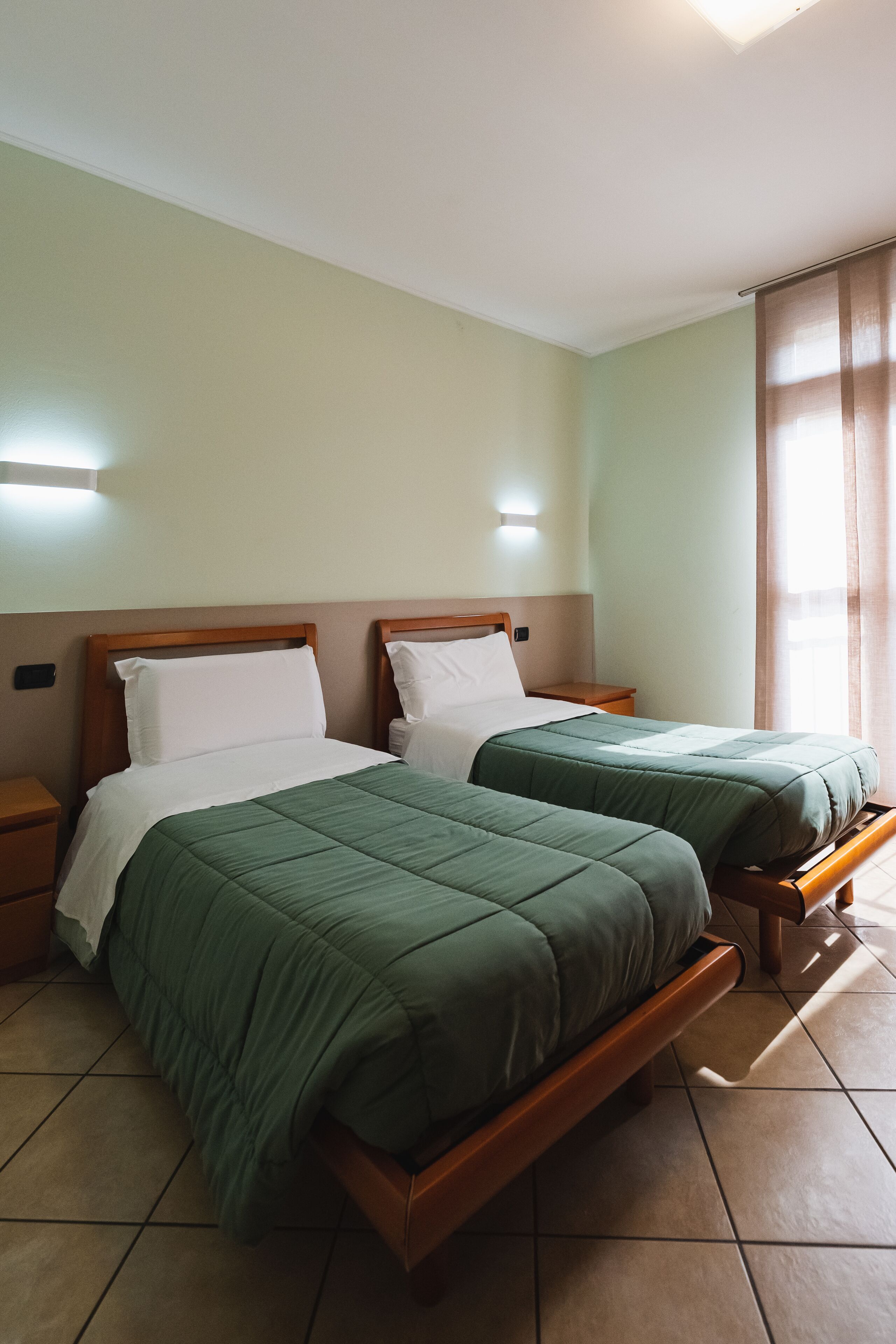 Habitación de hotel con dos camas individuales, cabeceros de madera y una mesita de noche con lámpara.