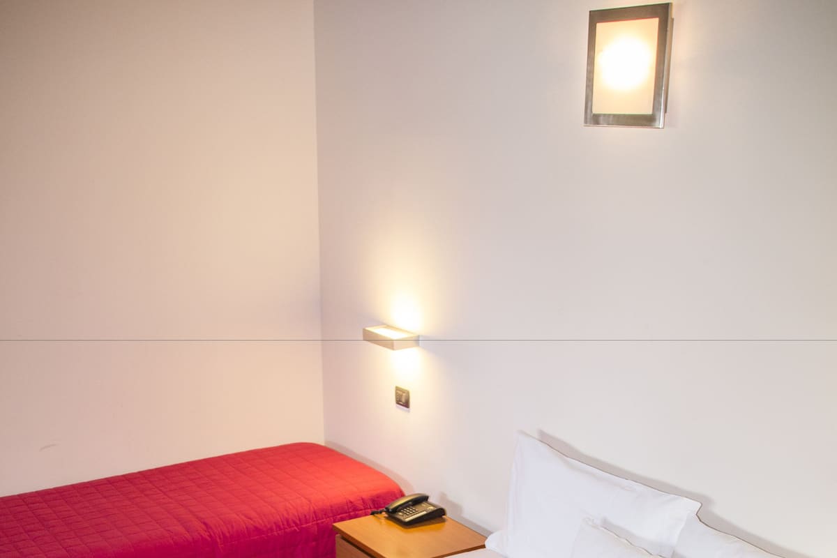 A hotel room with two beds, each with a red bedspread, a white headboard, and a bedside table with a lamp.