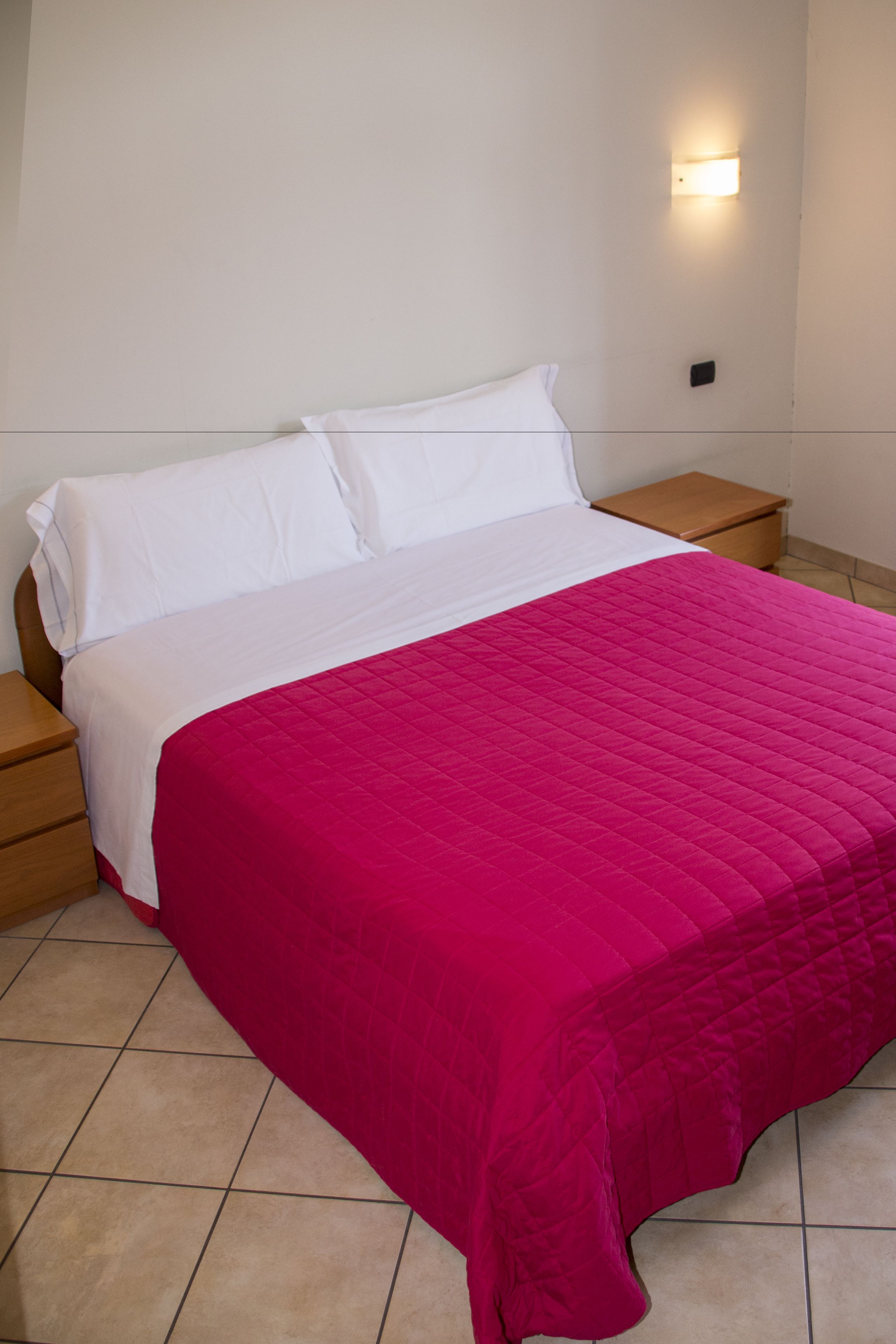 Un lit avec un matelas blanc et une couvre-lit rose matelassée, flanqué de tables de chevet en bois dotées d’un petit tiroir, dans une pièce aux murs unis et avec un luminaire placé au-dessus du lit.