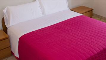 Un lit avec un matelas blanc et une couvre-lit rose matelassée, flanqué de tables de chevet en bois dotées d’un petit tiroir, dans une pièce aux murs unis et avec un luminaire placé au-dessus du lit.