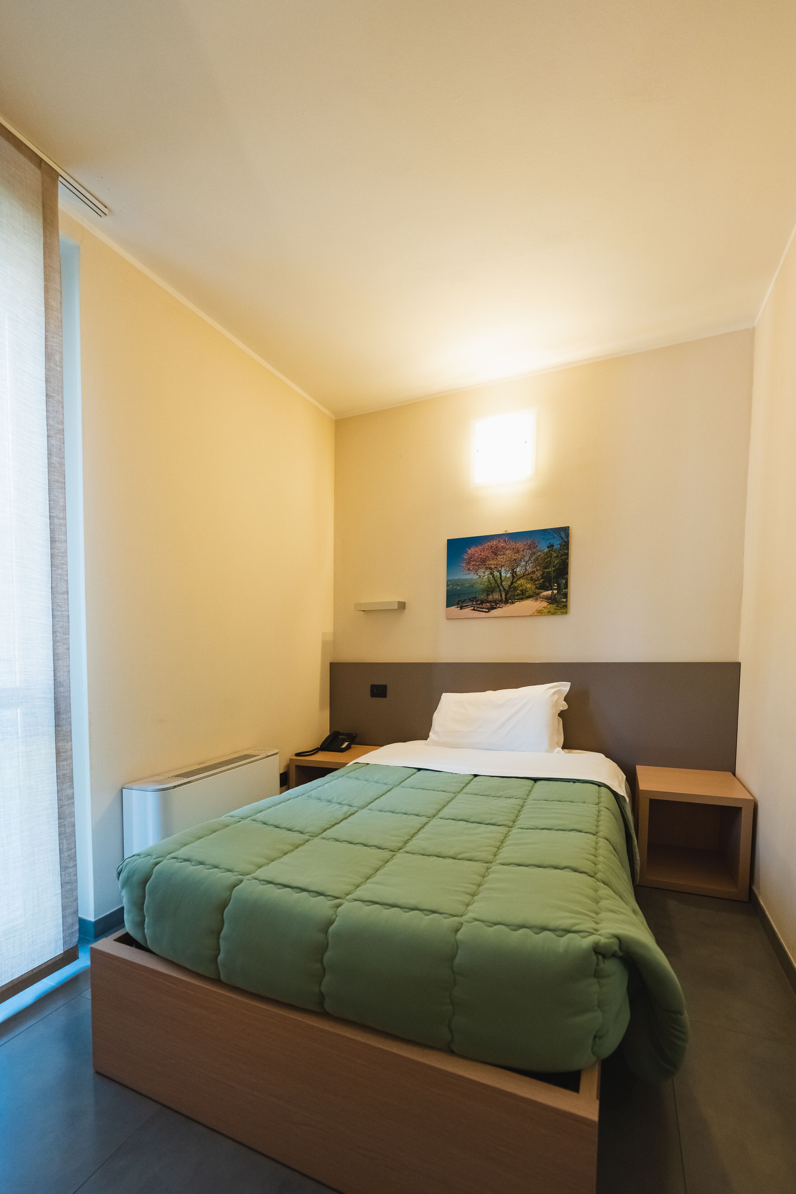 A hotel room with a bed, bedside tables, a nightstand, a painting on the wall, and a window with curtains.