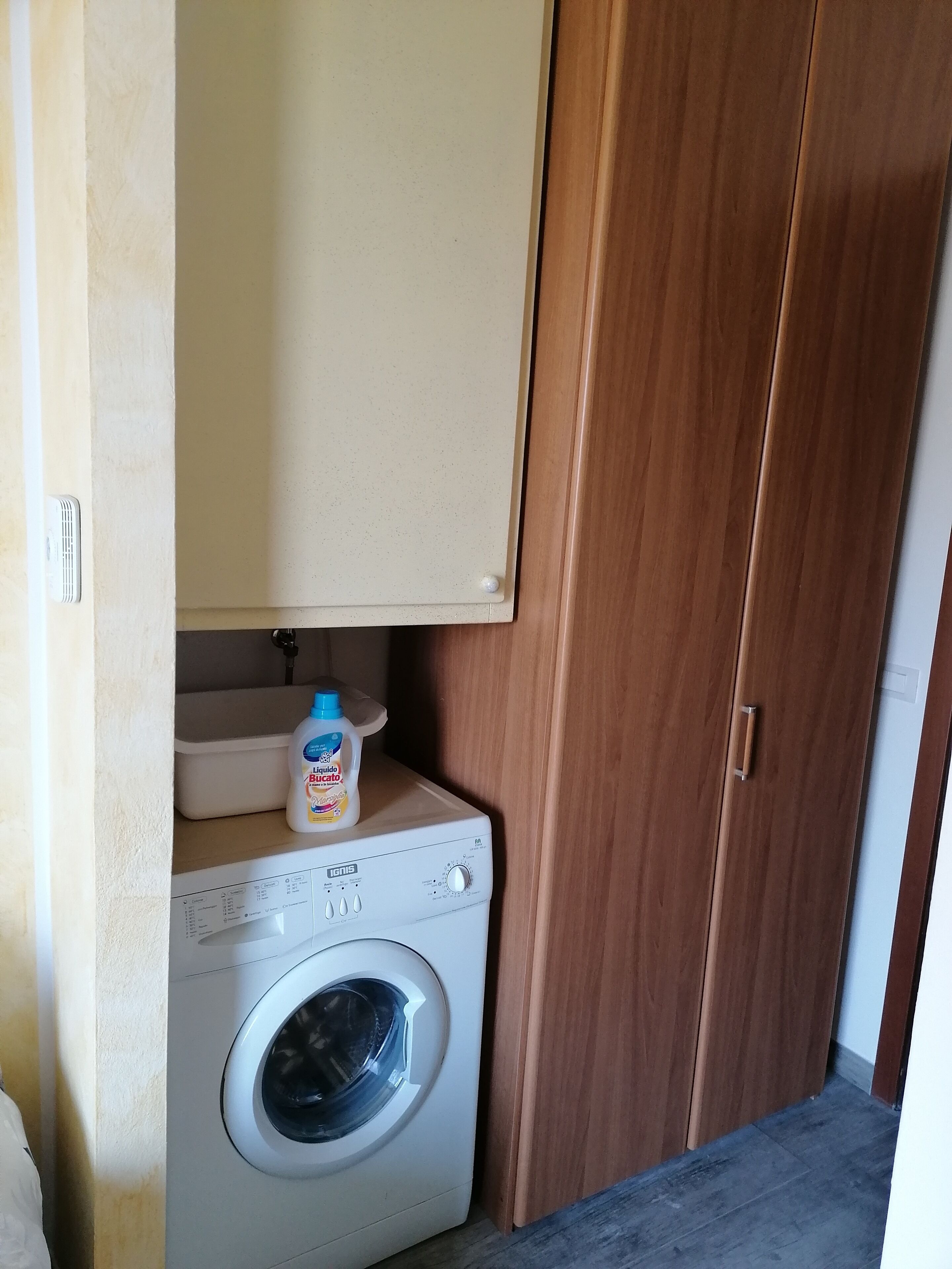 Una lavadora compacta con panel de control, una botella de detergente y un armario de madera en un pequeño lavadero.