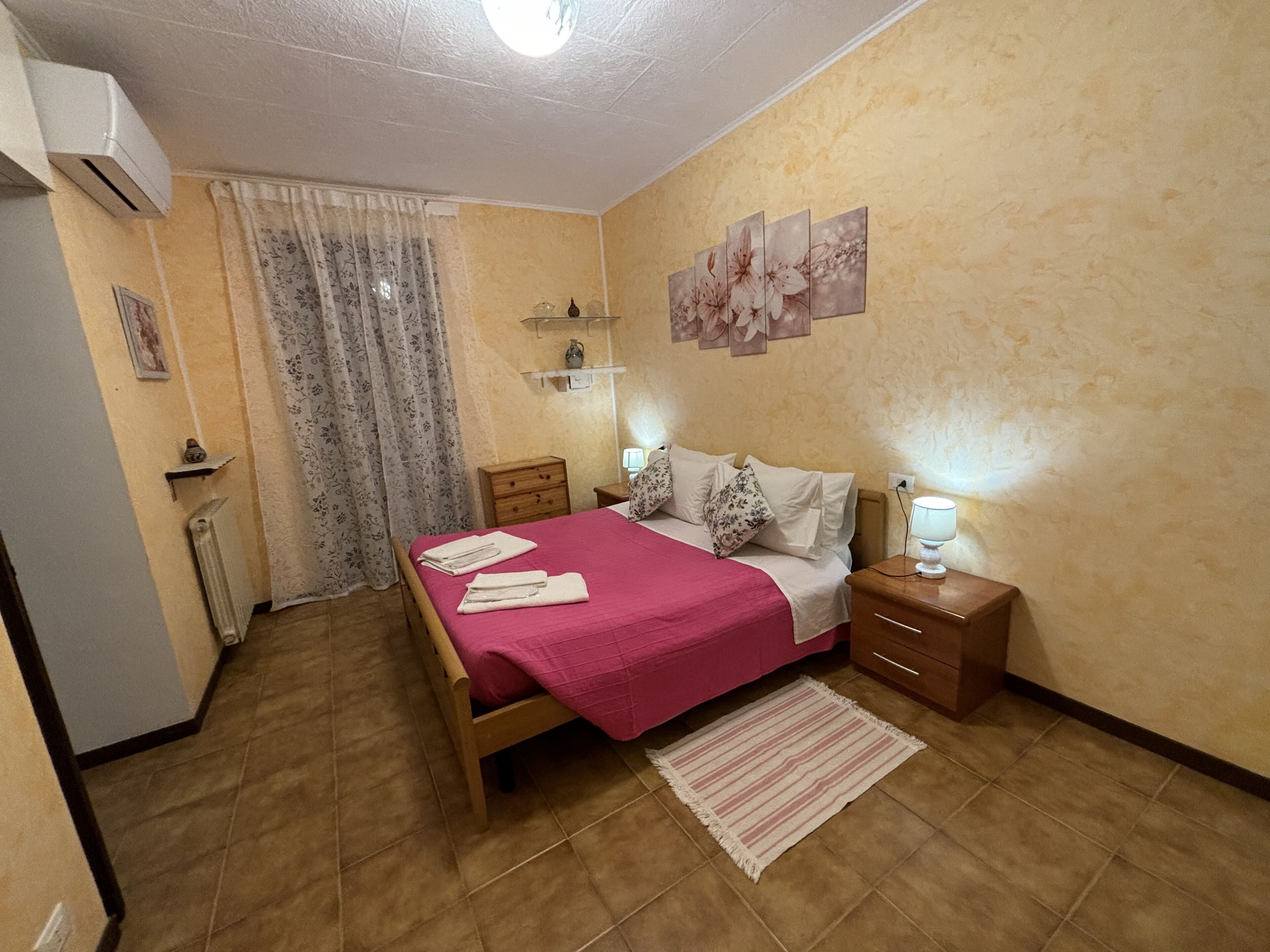 A bedroom with a bed, nightstands, a radiator, a window with curtains, and wall art.
