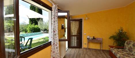 Habitación con piso de madera, sofá, una mesita con planta y vista a una piscina y vegetación.