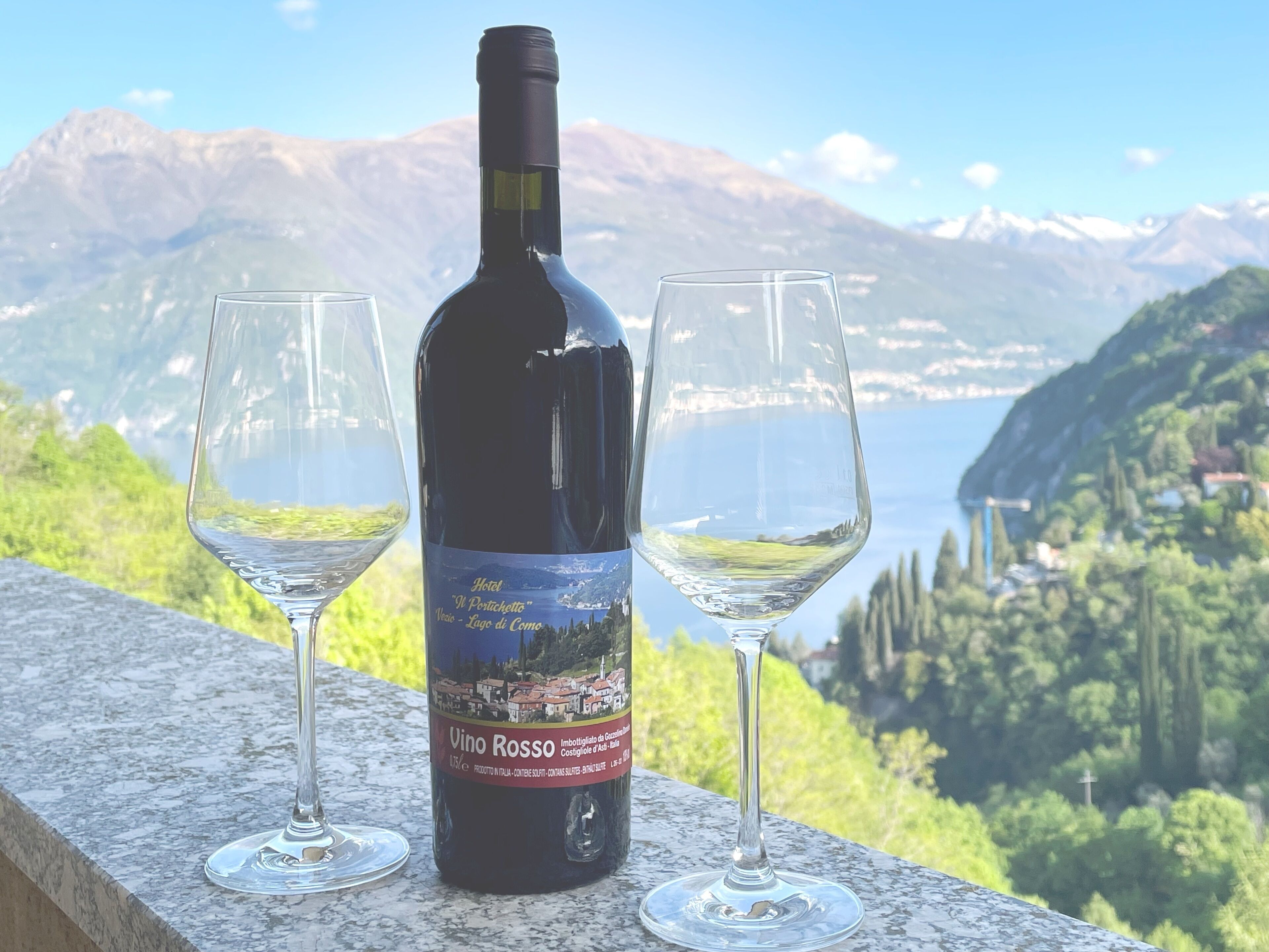 A bottle of Vino Rosso wine with two empty wine glasses on a ledge, overlooking a mountainous landscape with a lake.