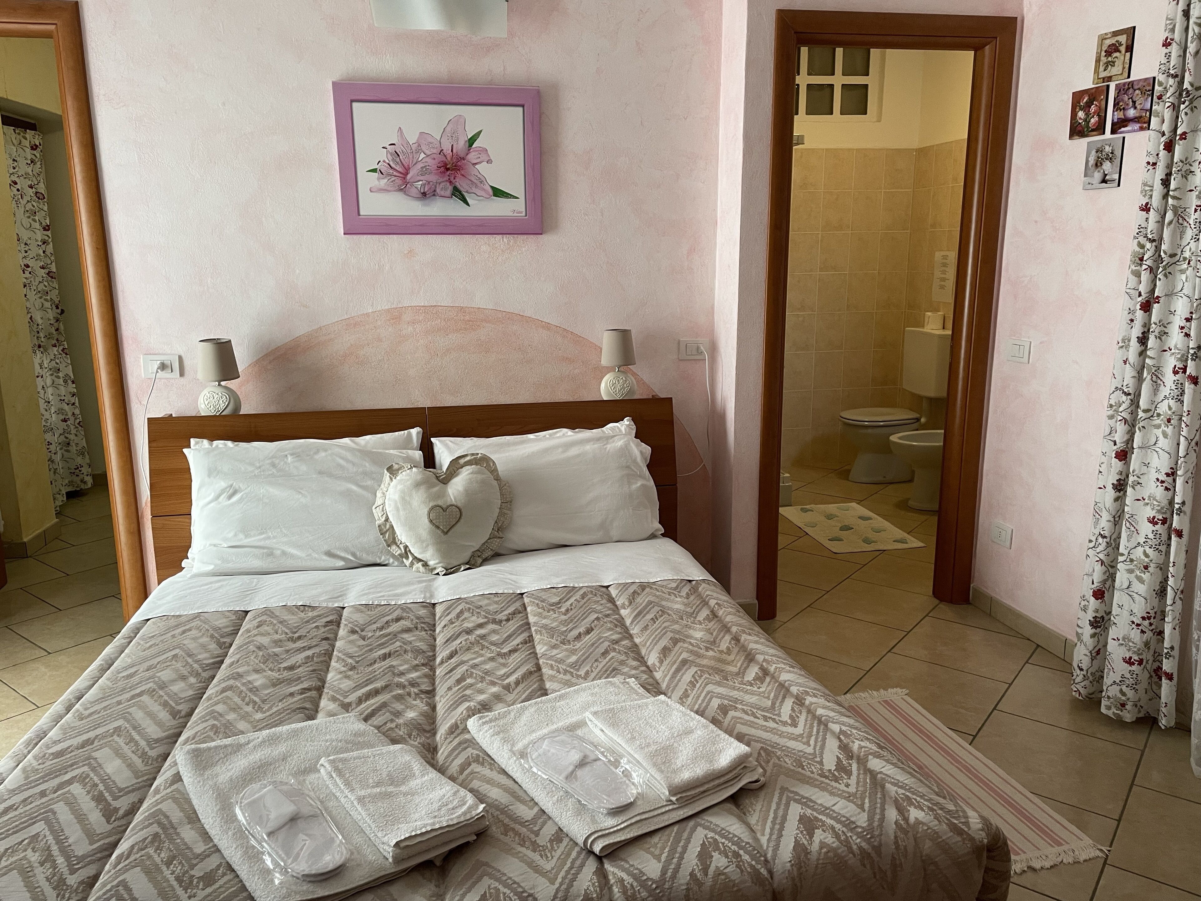 Ein ordentlich bezogenes Bett mit einem herzförmigen Kopfkissen, ein gerahmtes Blumenbild an der Wand und ein durchgehender Türspalt zum Badezimmer.
