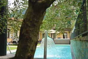 A swimming pool with a water feature, surrounded by greenery and a stone wall. - Hotel Diffuso Il Portichetto sulla collina a Vezio Perledo (Perledo)