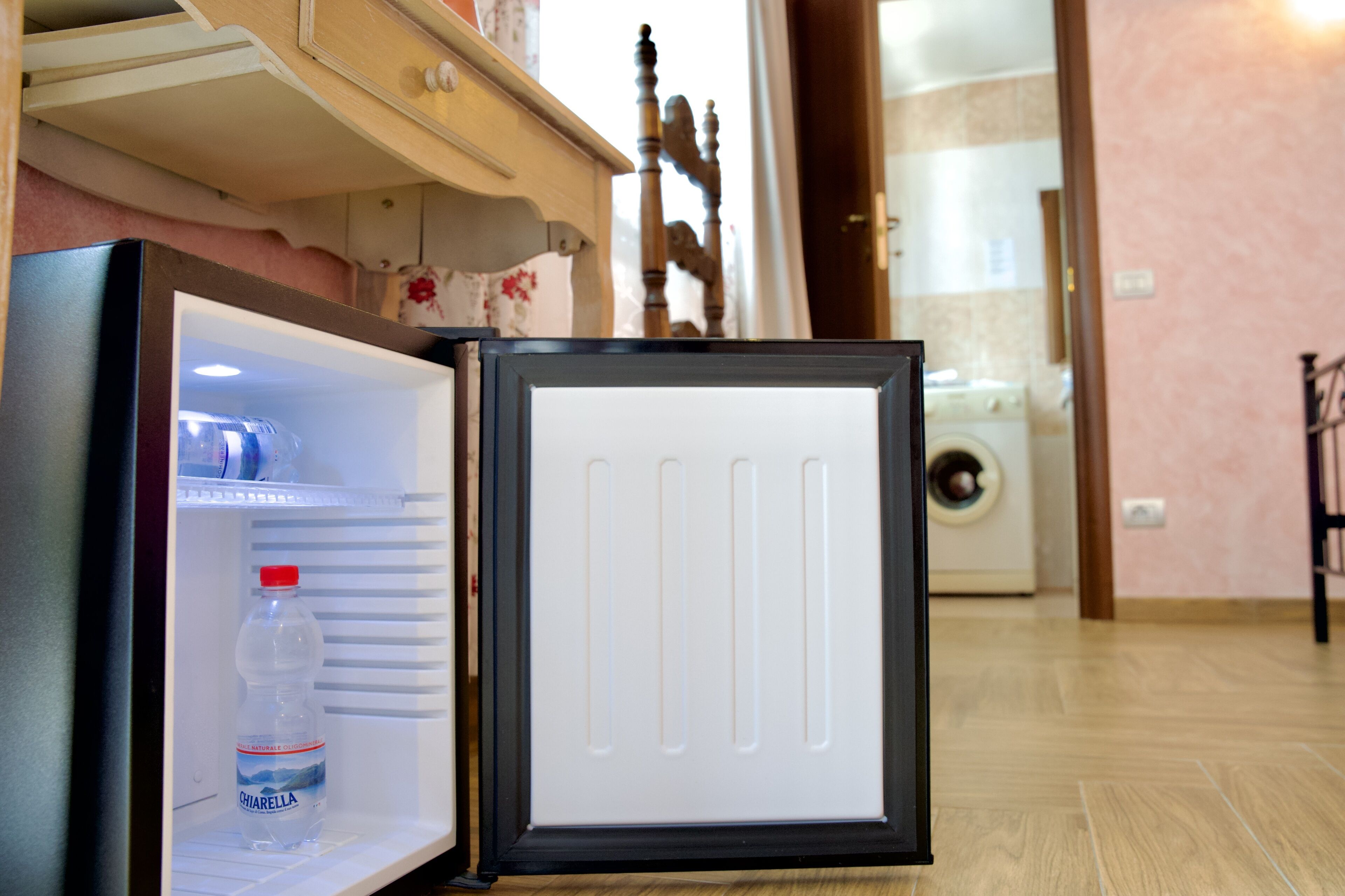 Un refrigerador compacto con una botella de agua Chiarella en su interior, una silla de madera y una lavadora al fondo.