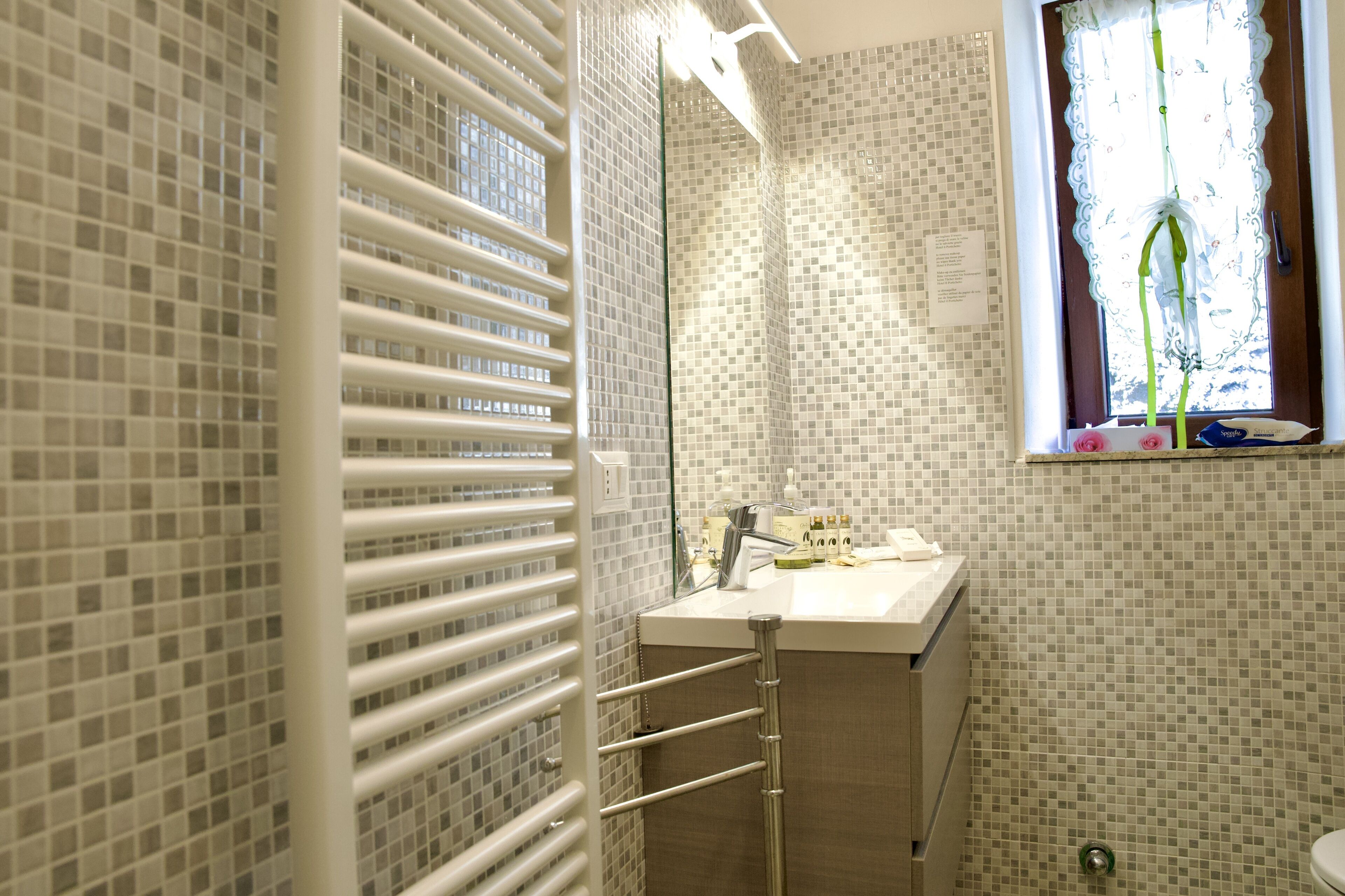 Un baño con pared de azulejos, un lavamanos blanco, un espejo y una ventana con cortina.