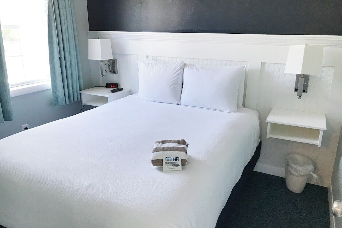 A hotel room with a large bed, white bedding, a bedside table with a lamp, a nightstand, and a small shelf.