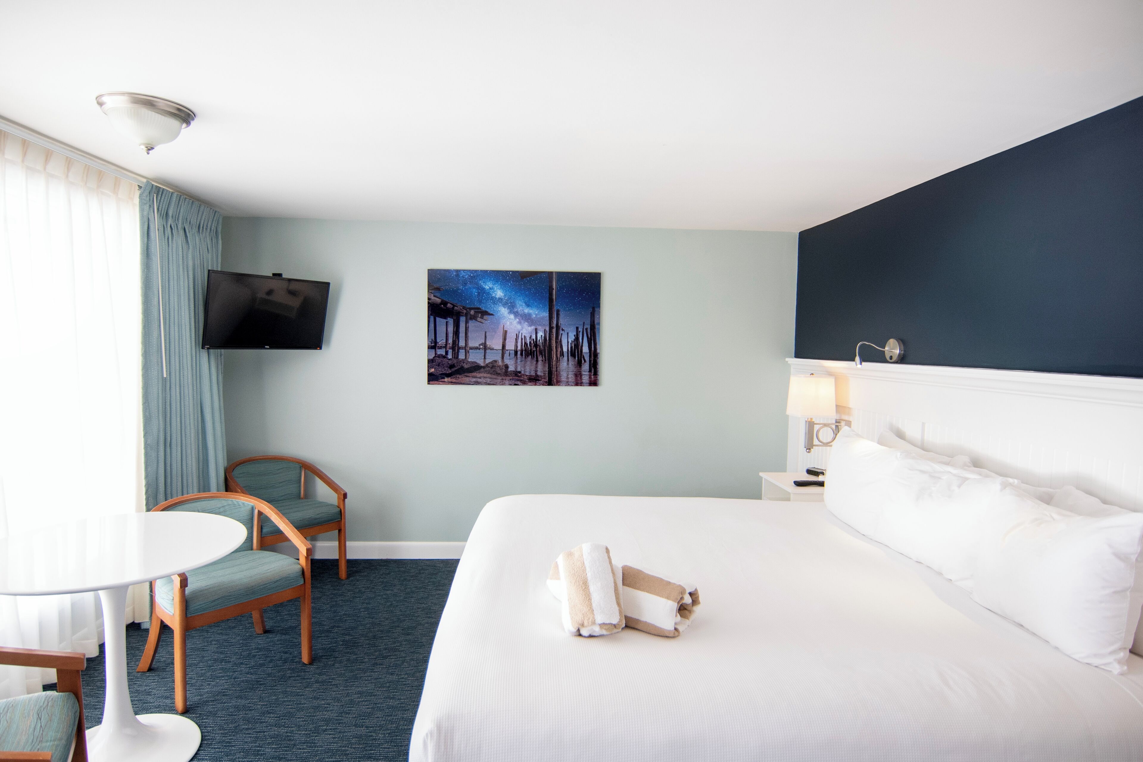 A hotel room with a bed, a TV mounted on the wall, a painting of a night sky, a desk with a lamp, and two chairs.