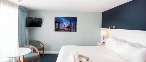 Une chambre d’hôtel avec un lit, un téléviseur fixé au mur, un tableau représentant un ciel nocturne, un bureau avec une lampe et deux chaises.