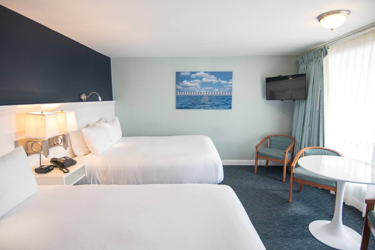 A hotel room with two beds, a desk with a lamp, a chair, a TV mounted on the wall, and a painting of a bridge over water.