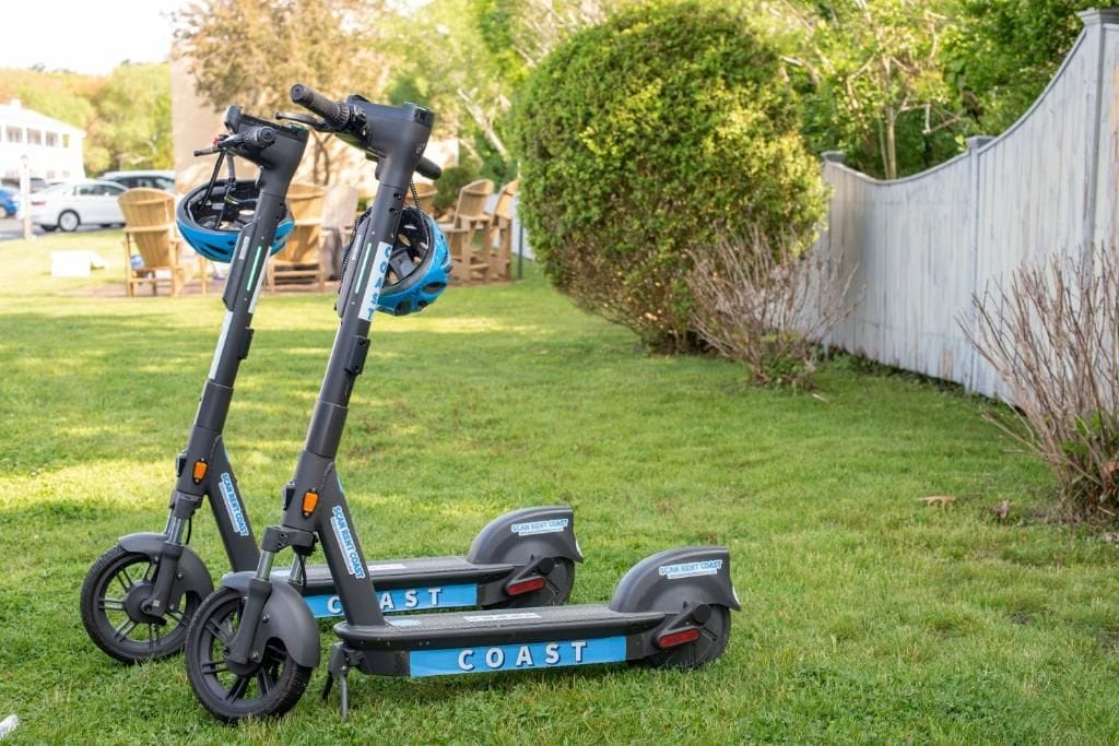 Two electric scooters on a grassy lawn with a white fence and greenery in the background.