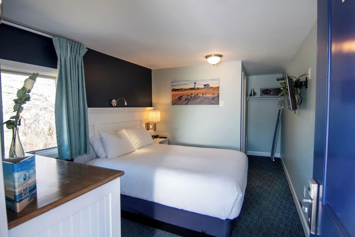 A hotel room with a bed, a nightstand, a lamp, a picture on the wall, and a window with curtains.