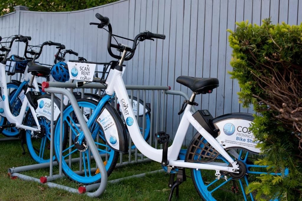 A row of white and blue bicycles with 'COAST' branding, parked against a grey fence.
