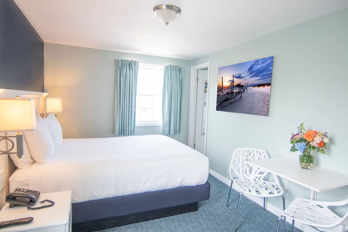 A hotel room with a bed, a desk, a chair, a lamp, a phone, and a painting on the wall.