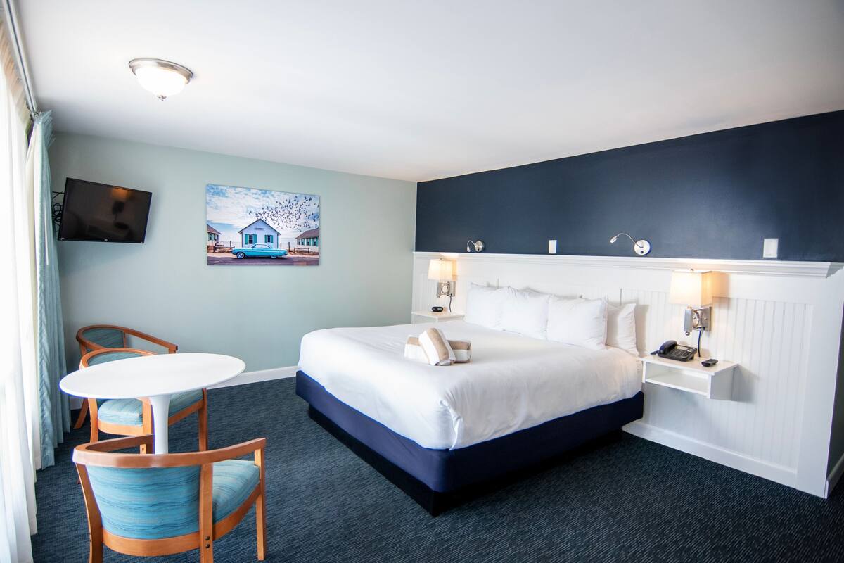 A hotel room with a bed, a desk, two chairs, a television, and a painting on the wall.