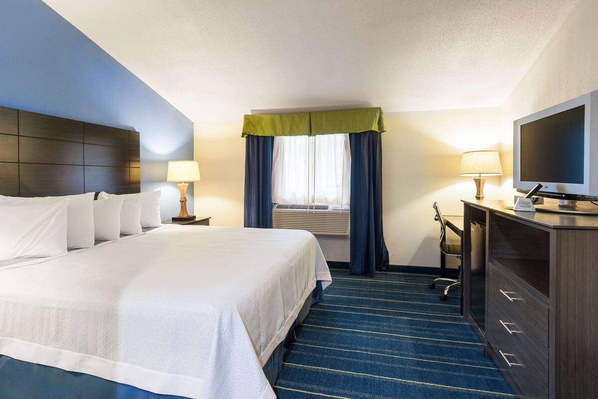 A hotel room with a large bed, a desk with a television, a chair, and a window with curtains.