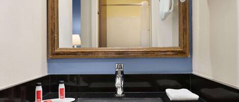 Une salle de bain avec un plan de toilette noir, un lavabo blanc, un miroir avec un cadre en bois et deux bouteilles de shampoing.