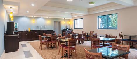 A spacious dining area with multiple tables and chairs, a kitchen in the background, and large windows allowing natural light.