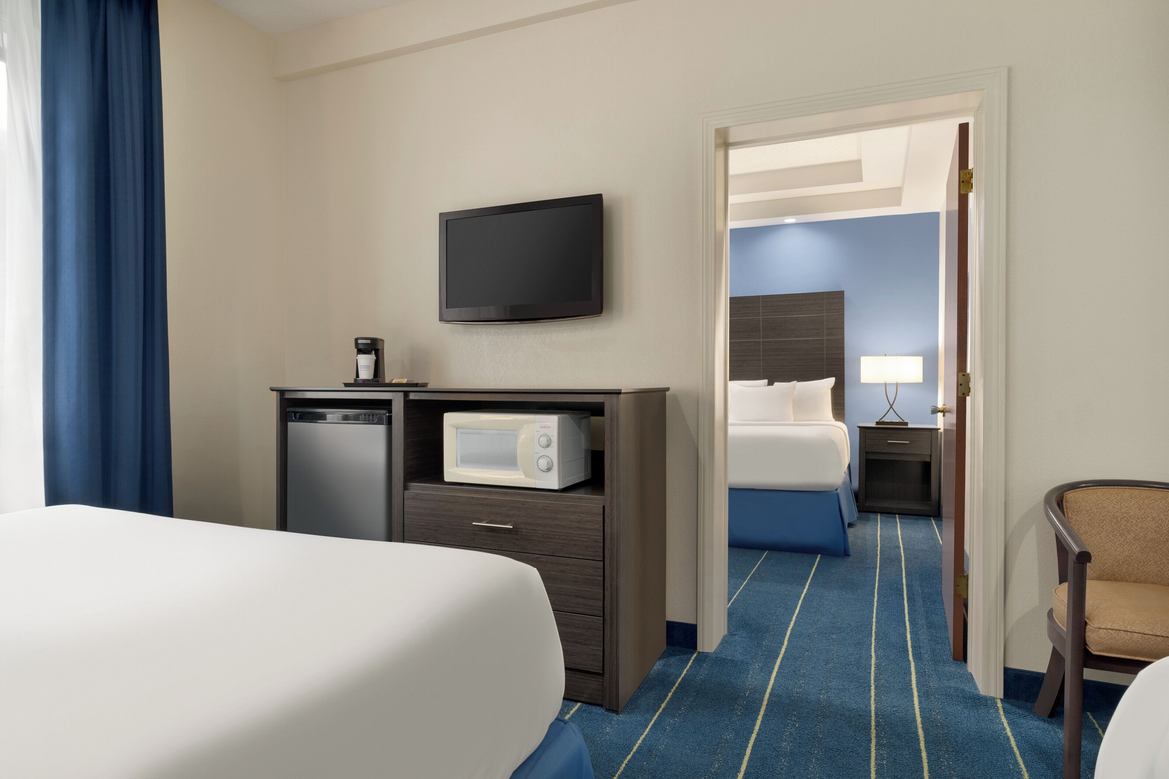 Quarto de hotel com cama, escrivaninha com micro-ondas, televisão e mesa de cabeceira com luminária.