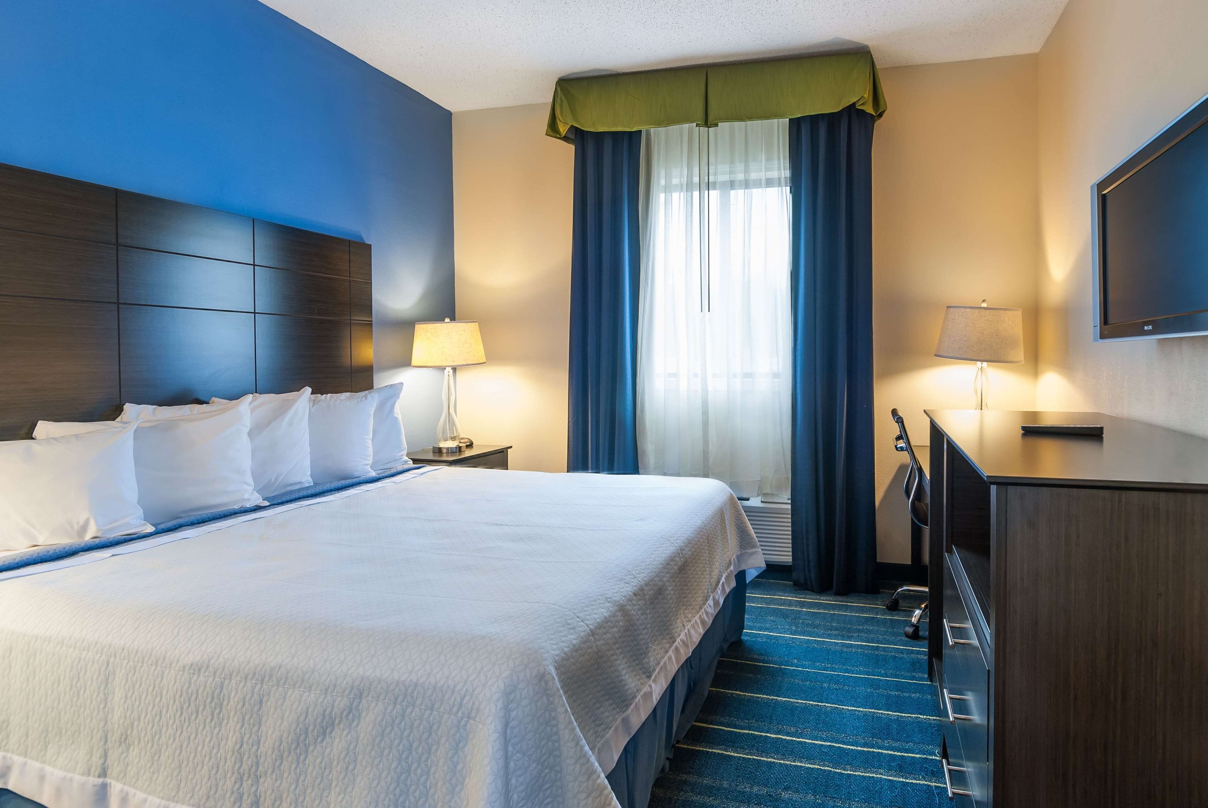 A hotel room with a large bed, two bedside tables with lamps, a desk with a chair, and a flat-screen TV on the wall.