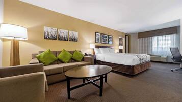 A hotel room with a bed, a sofa with green cushions, a coffee table, a desk with a lamp, and framed pictures on the wall.