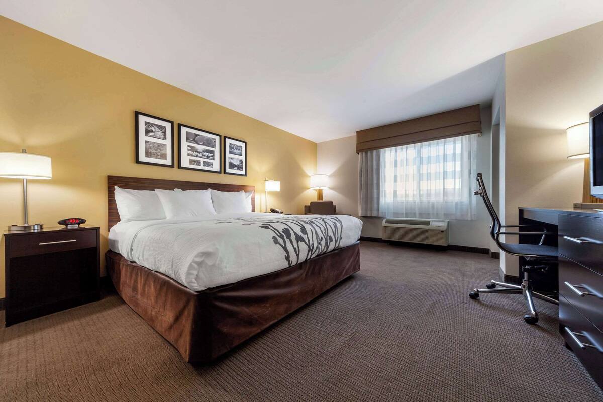 A hotel room with a large bed, bedside tables, a desk, and a chair. There are framed pictures on the wall, a window with curtains, and an air conditioning unit.