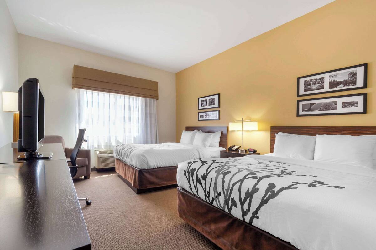 A hotel room with two beds, a desk with a television, a chair, and a window with curtains. There are framed pictures on the wall.