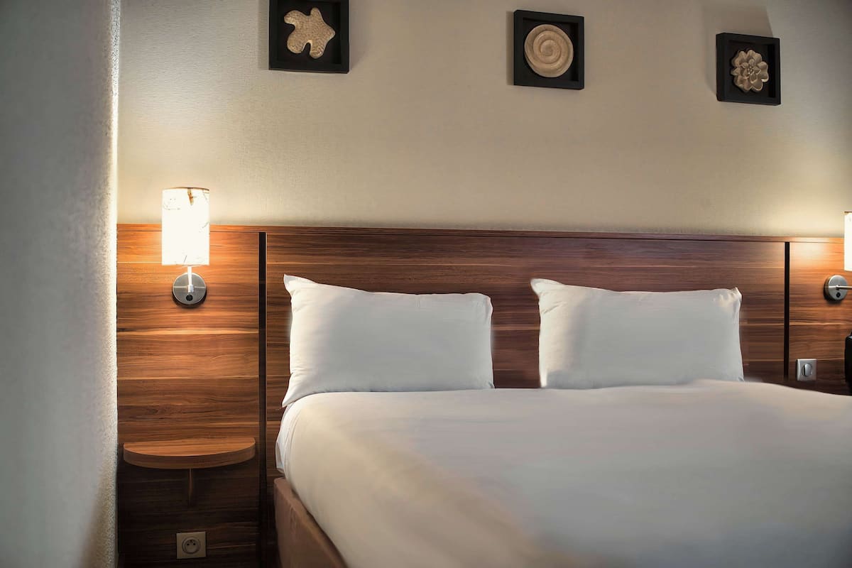 Un lit bien fait, avec du linge de lit blanc, une tête de lit en bois et deux tables de chevet fixées au mur, chacune surmontée d’une lampe.
