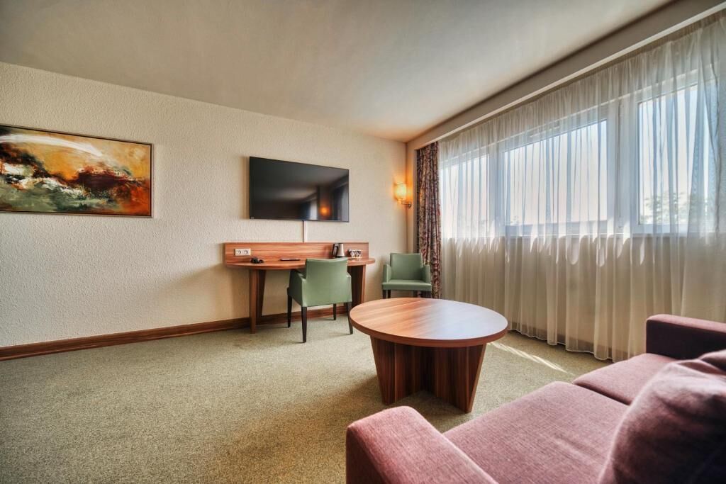 Habitación de hotel con escritorio de madera, dos sillas, una mesa de centro redonda, televisor de pantalla plana montado en la pared y un amplio ventanal con cortinas de encaje.