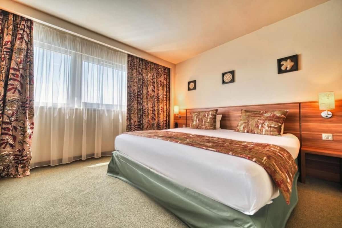 Une chambre d’hôtel avec un grand lit, une tête de lit en bois, deux tables de chevet avec des lampes et une fenêtre avec des rideaux légers.
