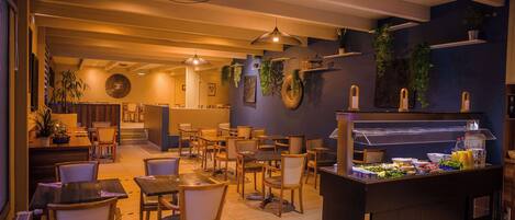 A restaurant with multiple dining tables and chairs, a buffet counter with food items, and hanging plants on the walls.