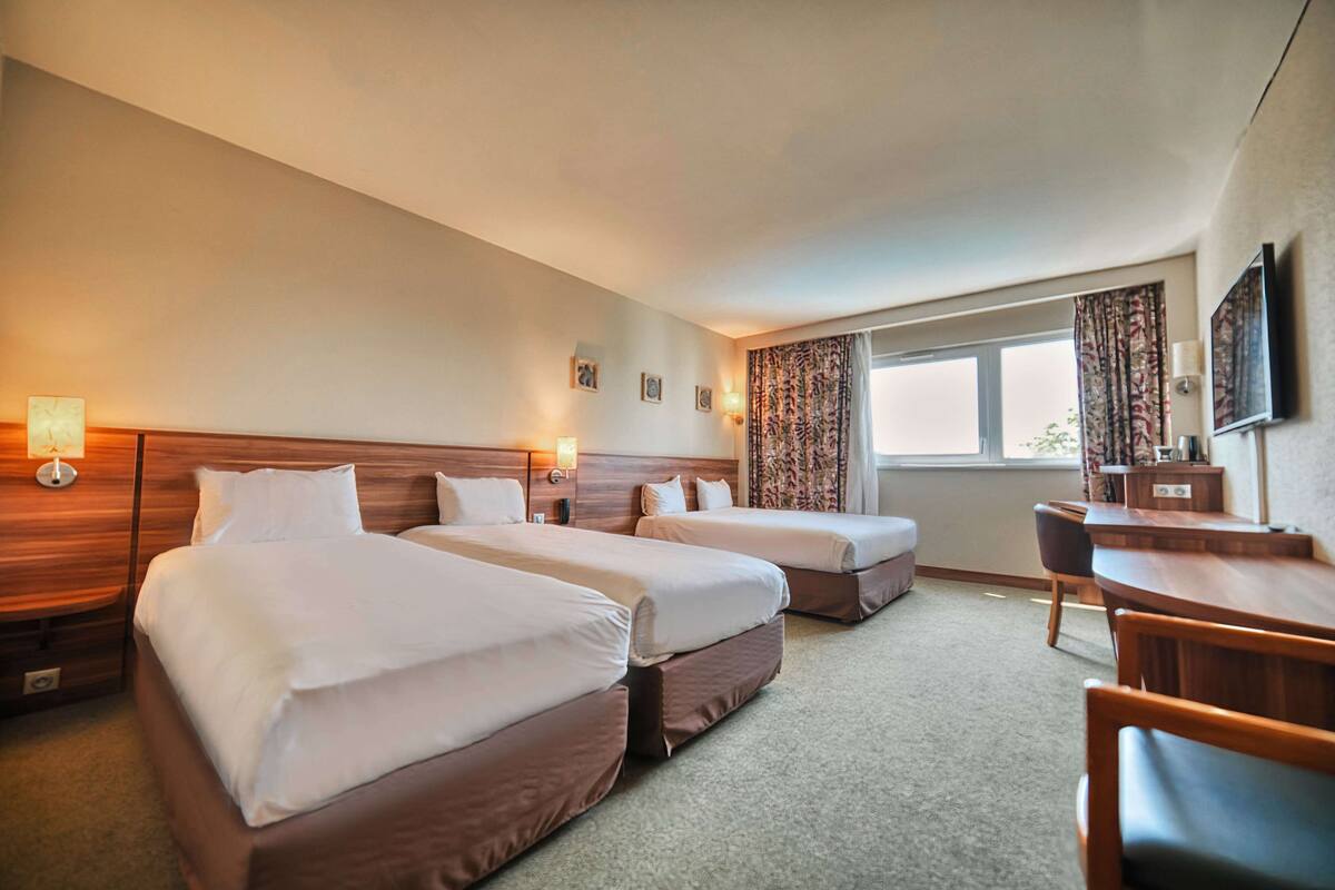 Une chambre d’hôtel avec deux lits, un bureau et une chaise. On y trouve des lampes fixées au mur et une fenêtre avec des rideaux.