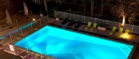 A rectangular swimming pool with a tiled deck and a metal fence. There are lounge chairs and umbrellas along the poolside.